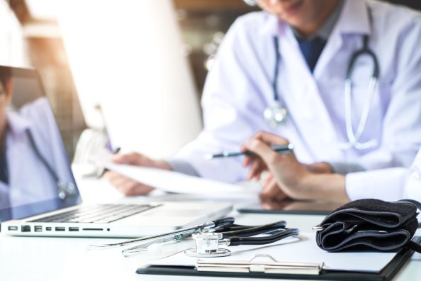 Two doctors being discussing patient history in an office pointing to a clipboard with document paper as they make a diagnosis or recommend on treatment